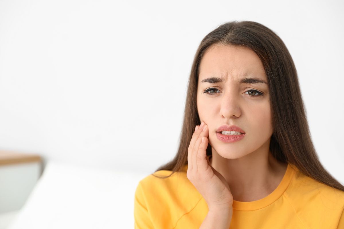 Woman in yellow shirt holding her cheek, showing discomfort from swollen gums near wisdom teeth.